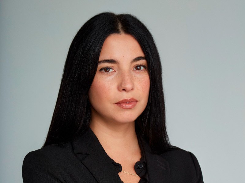 A woman with long black hair wearing a black shirt looks at the camera in front of a grey background.