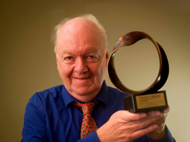 A man wearing a blue shirt and red tie holds a bronze-coloured award up. He is Prof Colm Mulcahy.