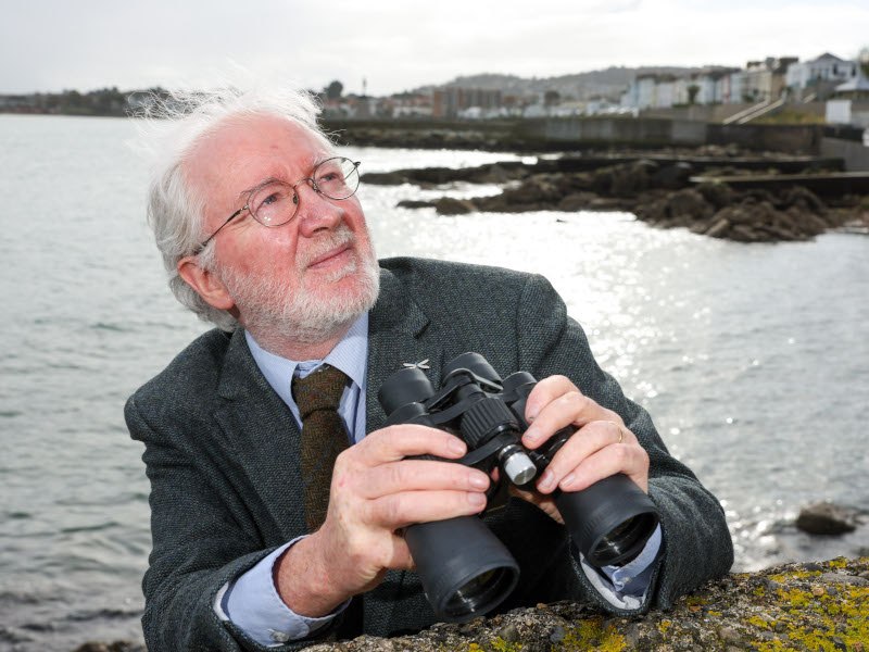 A man with white hair and a beard leaning against a wall looking up. He’s holding a pair of binoculars and a beautiful coastal scene is behind him.