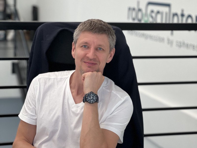 A photo of Dennis Ledenkof in a white t-shirt sitting at a desk in a high-backed chair. He is resting his head on his hand.