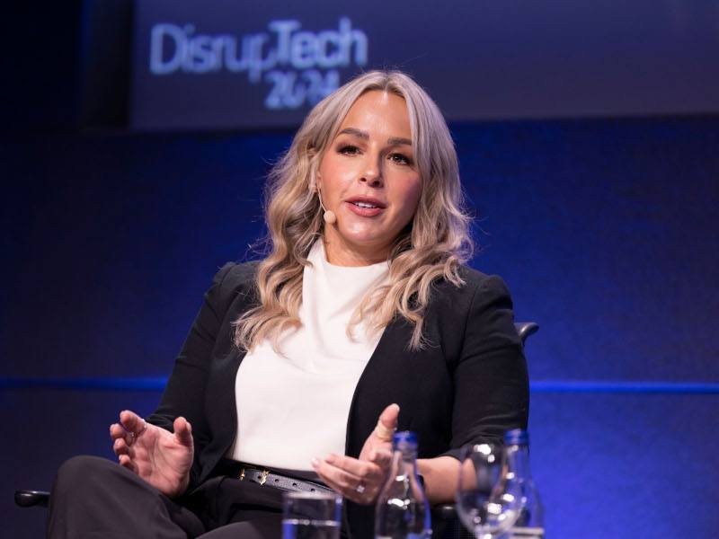 A woman with blonde hair sits on a stage with a hands-free microphone talking. A sign behind her says DisrupTech 2024.