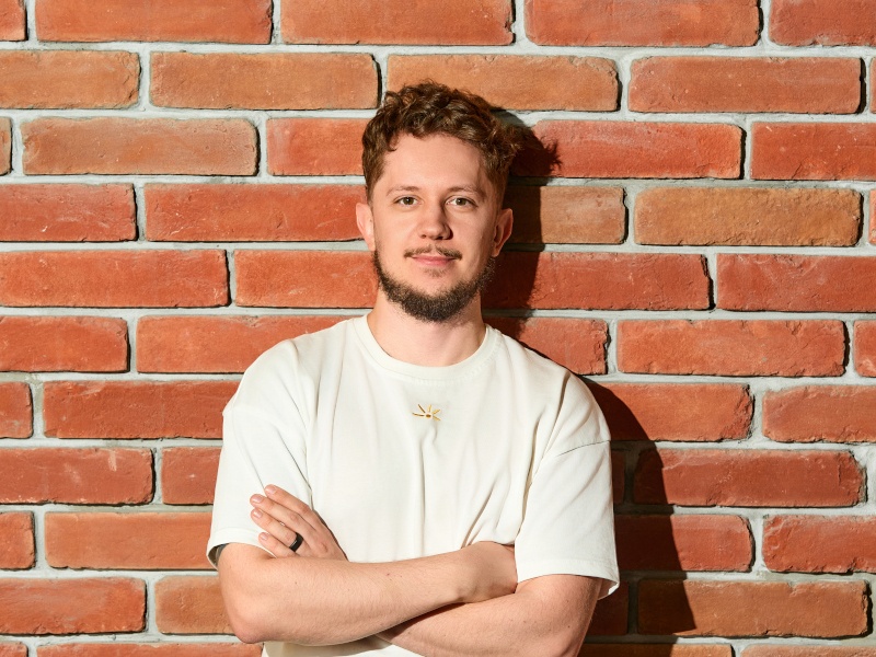 A bearded man wearing a white T-shirt smiles at the camera with his arms folded, while standing in front of a red brick wall.