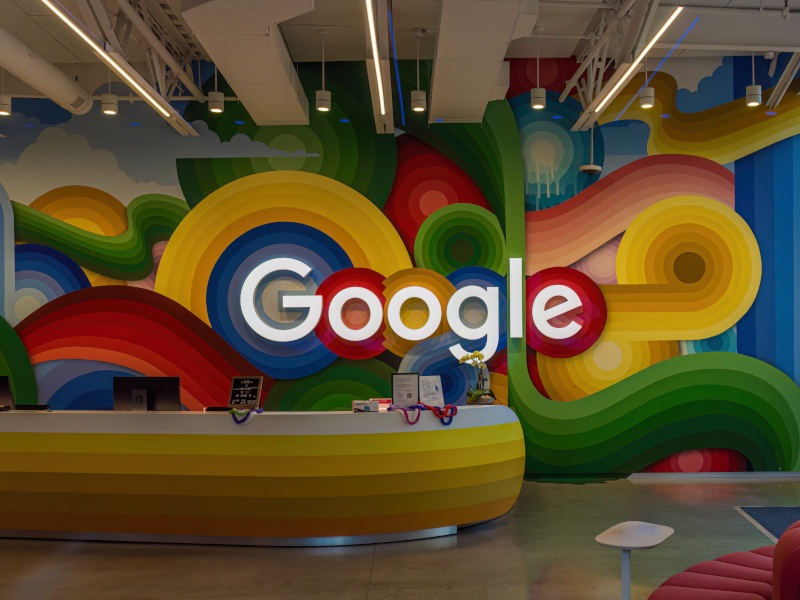 The Google logo with colourful outlines on a wall behind a yellow reception desk inside an office building.