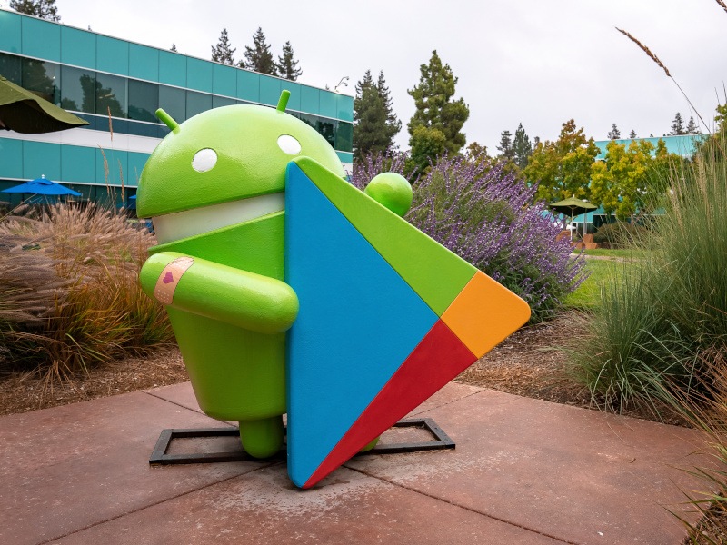 A statue of the green Android mascot holding a Google Play Store triangle logo in a garden. There is a glass building in the background.