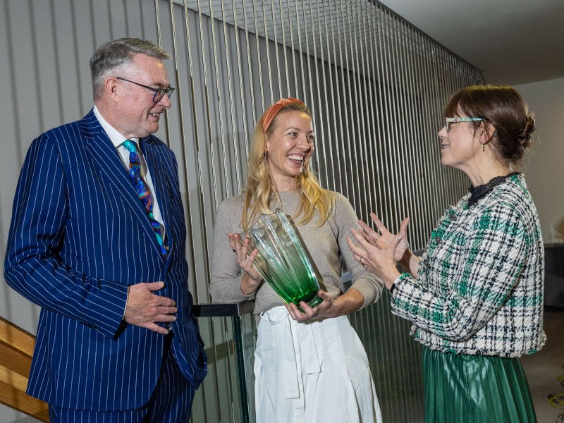 A tall man in a blue suit to the left, one women in the centre holding an award and a woman to the right in a green dress.