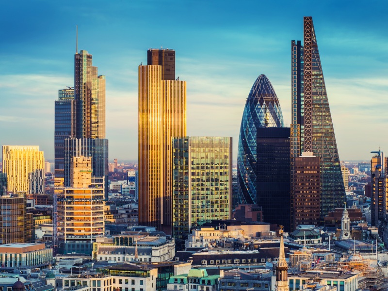 The London skyline during sunset. Tall buildings are reflecting sunlight with a blue sky in the back and many smaller buildings closer to ground.