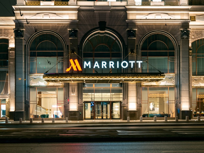 A lit-up Marriott logo sign on top of a large, decorative hotel with a road in front of it at night time.