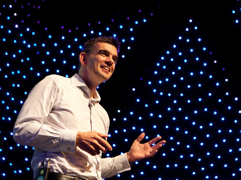 An upward-angled shot of a man with dark hair wearing a shirt and jeans talking on stage. Lots of tiny blue lights are on a screen behind him.