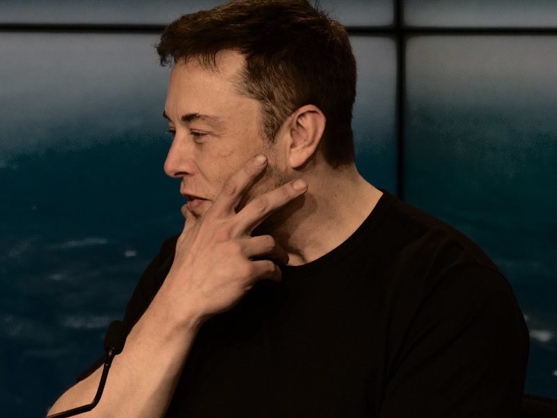 Elon Musk holds his chin while looking away from the camera.