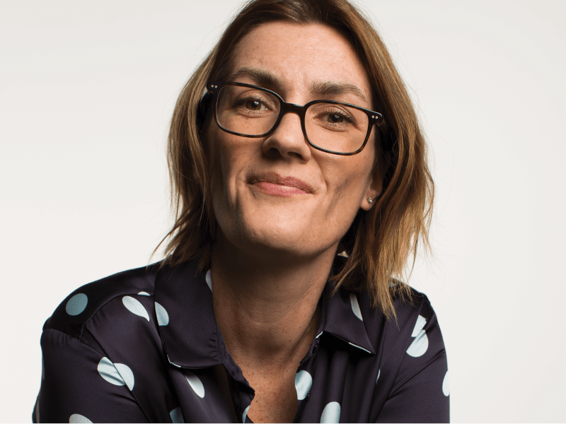 A woman wearing glasses and a dark shirt with white circular patterns smiles at the camera.