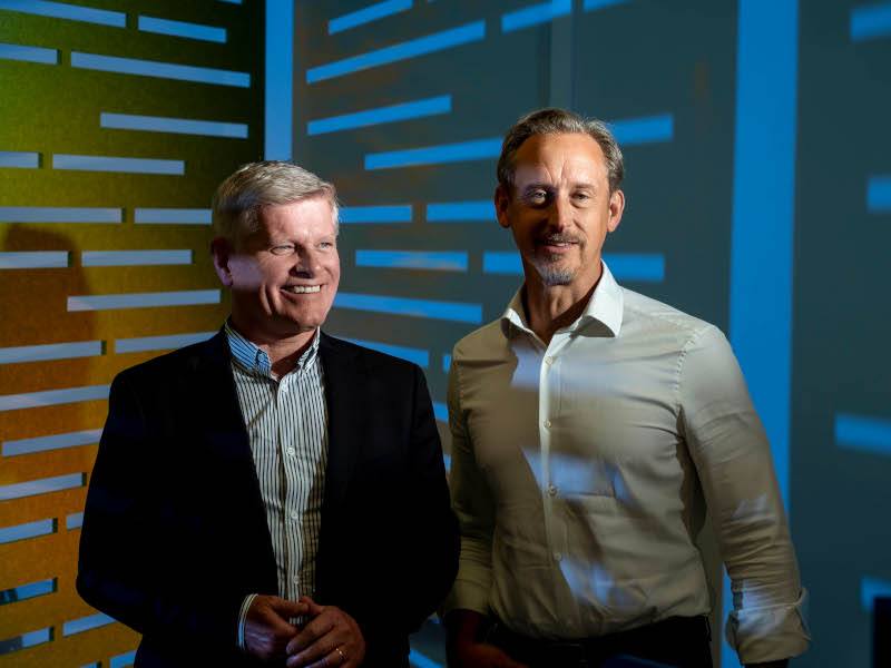 Two men stand side by side in a corner with blue walls. Lights cast shadows behind them. They are smiling at the camera.