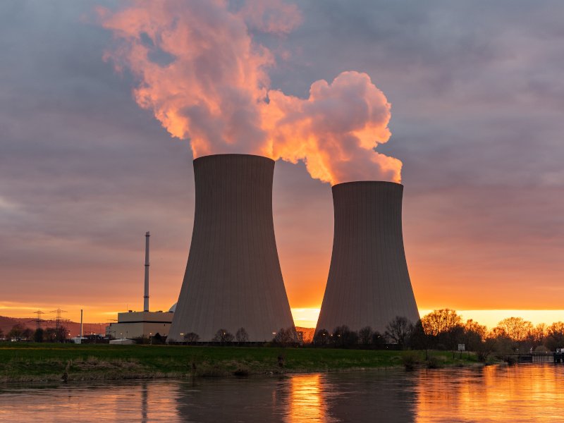 A nuclear power plant at the edge of a body of water against a sunset. Large plumes of smoke are coming out of the two cooling towers.