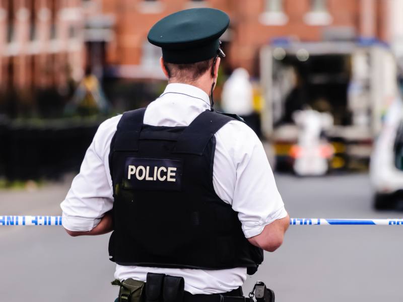 A PSNI officer with his back to the camera. wearing a black vest that says police on the back. He’s standing in front of blue and white police tape.