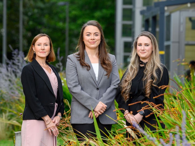 Three women standing outdoors with tall plants in the foreground.