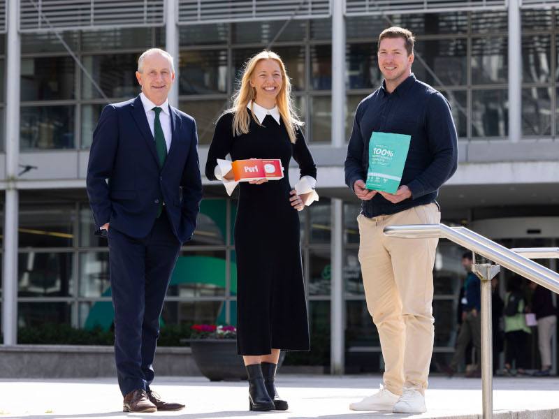 Three people standing outside. The man to the left has his hands in his pockets, while the person in the middle, a woman and a man to her right are holding their products.