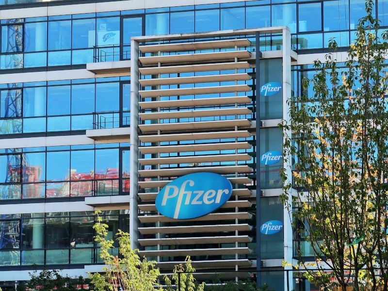 A large glass building on a sunny day with trees in front of it. It has a large Pfizer logo on the front with more in the windows.