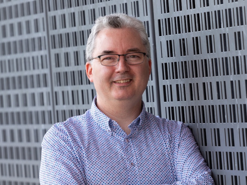 A man wearing a light-coloured shirt and glasses smiles at the camera.