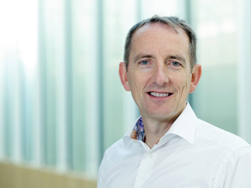 A headshot of Fergal Malone. He wears a white shirt and smiles at the camera in front of glass.