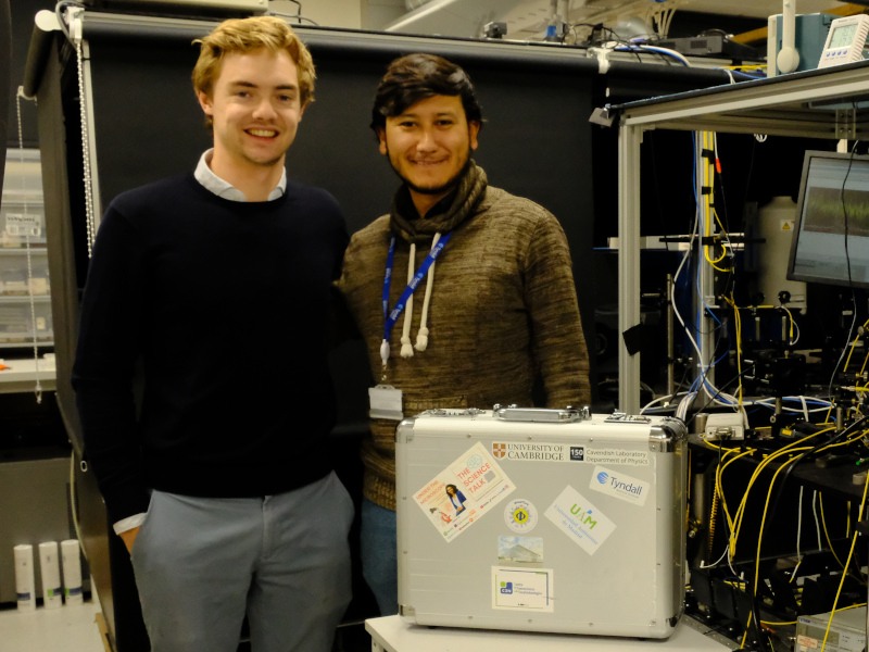 Two young men stand in a lab beside a silver suitcase with wires in the background.