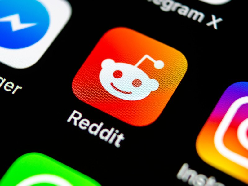 A close-up of the Reddit logo on a smartphone screen surrounded by other social media apps.