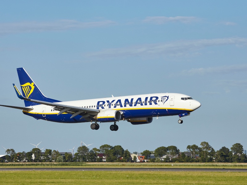 A Ryanair plane close to landing on the runway on a clear, sunny day. There are trees and wind turbines in the background.