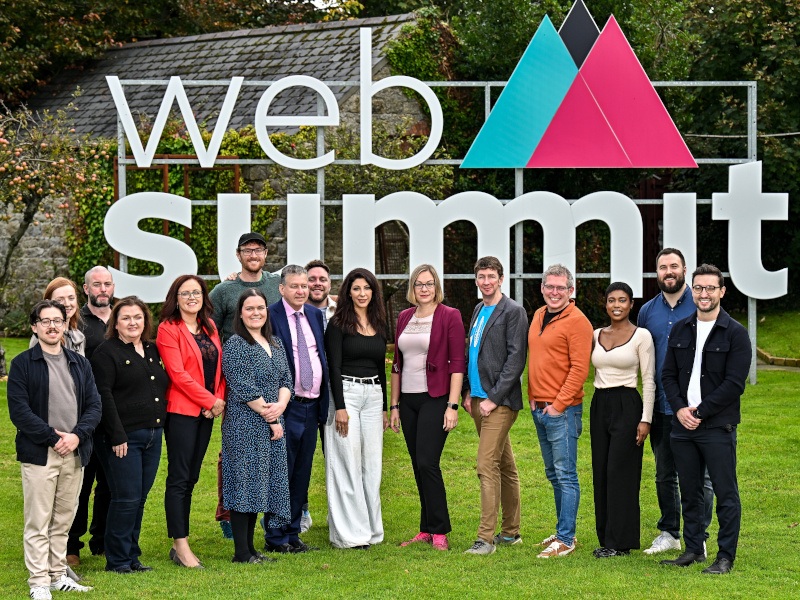 A group of men and women huddle on a green landscape in front of a large sign saying ‘Web Summit’.