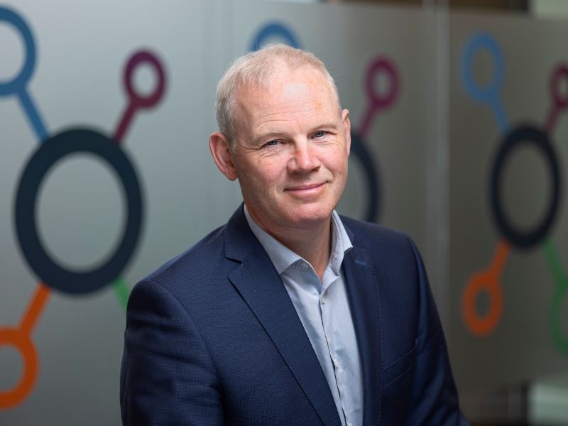 Headshot of a man wearing a navy coloured suit in an indoor space with the SuperNode logo on the wall behind him.