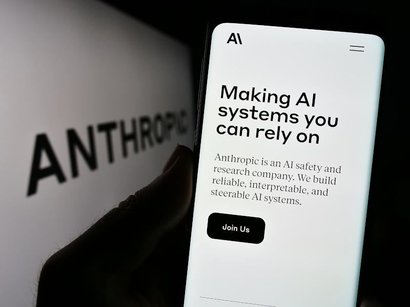 Person holding smartphone with web page of Anthropic. The screen reads "making AI systems you can rely on."