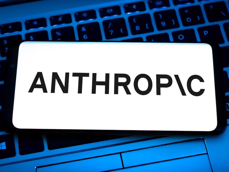 A laptop keyboard on the back with a blue hue and a smartphone with the Anthropic logo on the screen with a white background.