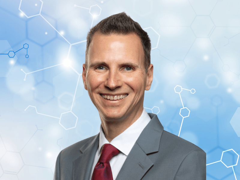 A headshot of David Henshall in a grey suit, red tie and white shirt against a pale blue abstract medical science background.