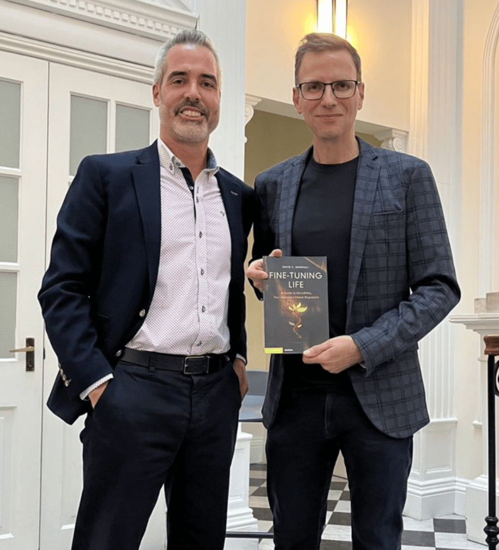 Kevin Mitchell and David Henshall stand next to each other in a large white room. David holds a copy of his new book, Fine-Tuning Life.