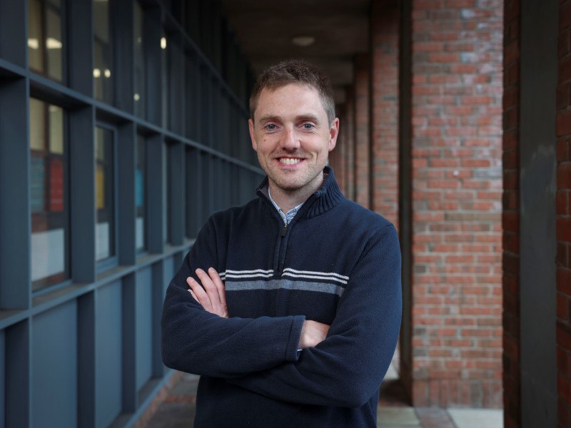 David Long stands outside in a stone walkway with his arms folded smiling at the camera.