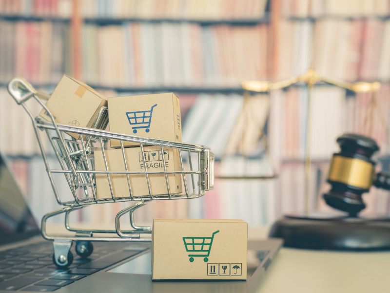 A blurry bookshelf in the background with a scale and a gavel to the right and a laptop with a small shopping cart and boxes to the left foreground.