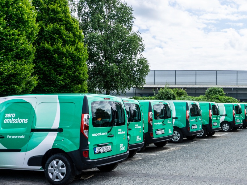 A row of electric, zero-emission delivery vans at an An Post Depot in Dublin during the day with trees in the back.