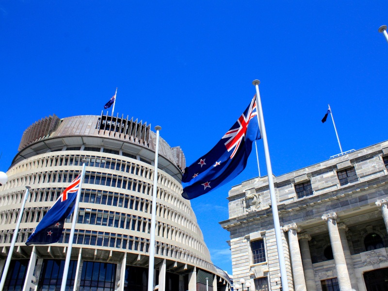 With a bright blue sky in the back and two New Zealand flags flying, this low angle shot showcases the country's parliament in Wellington city a slightly tapered cylindrical building.