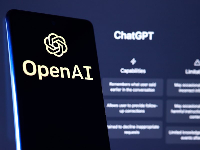 ChatGPT opened in a computer screen in the foreground. A smartphone with the OpenAI logo in the foreground to the left. The phone screen is black and the logo is in white.