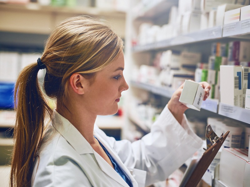 A female pharmacist is seen checking for medicine at a large shelf full of other medicines while holding a clipboard.