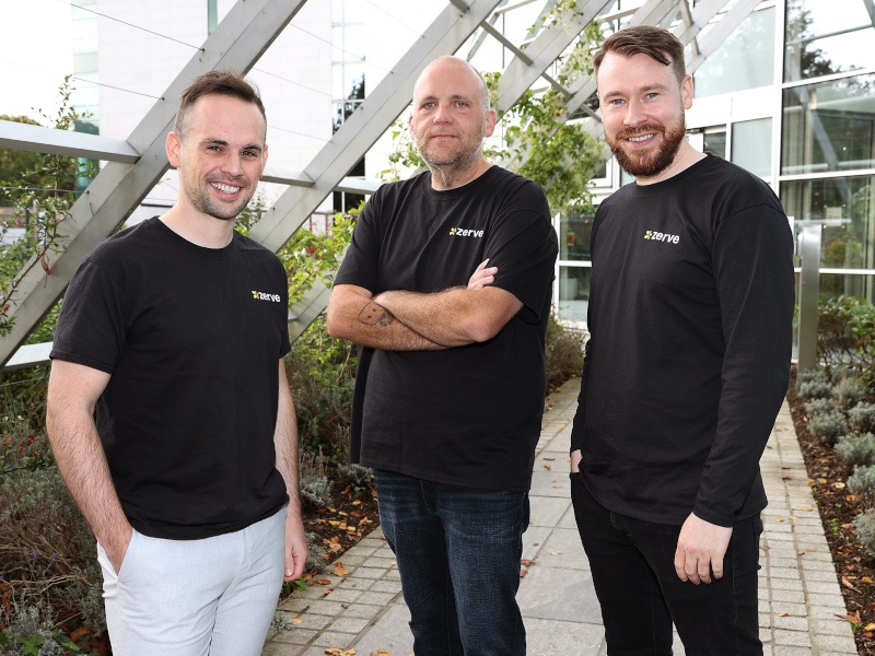 Three men standing outdoors, in front of a building on a cobblestone path with plants on both sides.