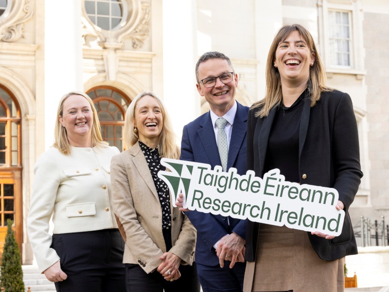 Four people standing in a group, with the lady in the front wearing a black blazer holding a a cut-out that says "Taighde Éireann – Research Ireland".