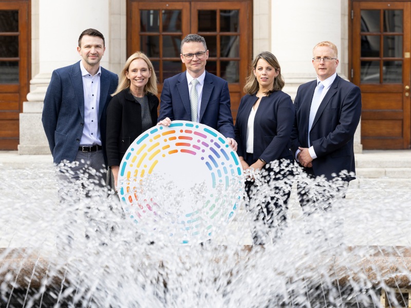 Five people standing behind a water fountain holding a circle cardboard cut out with colourful borders.