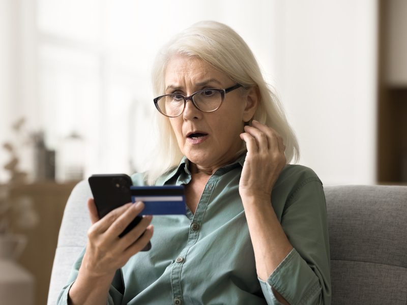 A person with white hair looking worried into their smartphone with an ATM card in their hand.