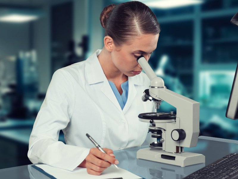 A female scientist in a lab examines something placed under a microscope. She is taking down notes of her observations onto a clipboard.