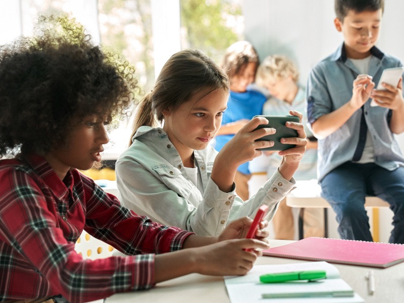 A small group of children are present in a classroom. They are all checking their phones. From a nearby window, it appears to be a sunny day outside.