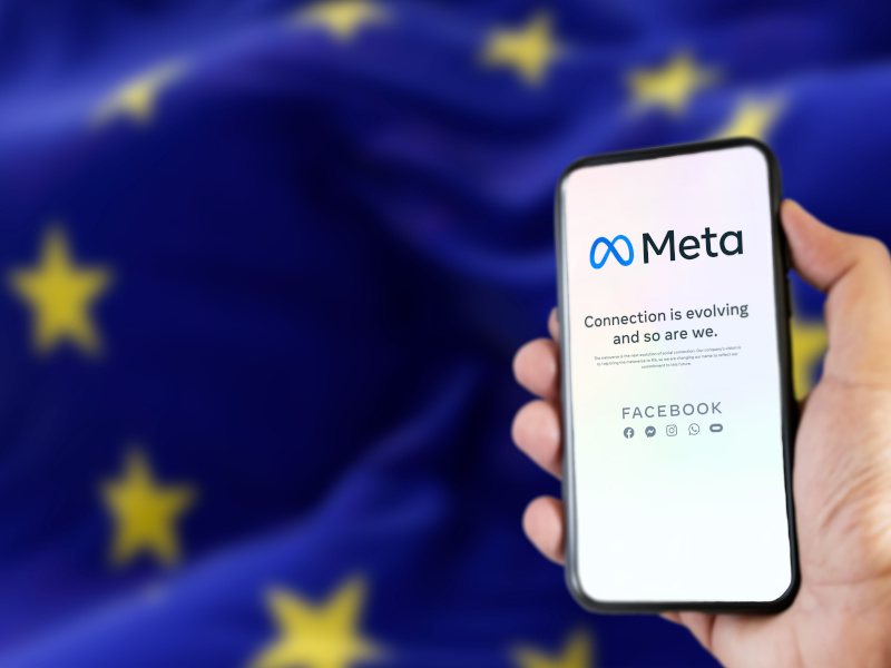 A smartphone is held in front of a blue flag with golden stars on it. The words ‘META’ along with the company’s logo are seen on its screen.