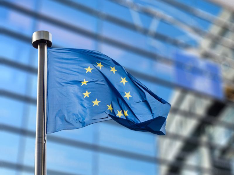 A waving blue flag adorned with a circle made up of stars flies outside of the official headquarters of the European Commission building.