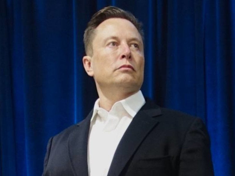 Entrepreneur Elon Musk, who is the richest person in the world. He is wearing a dark coloured suit and is standing against a blue background.