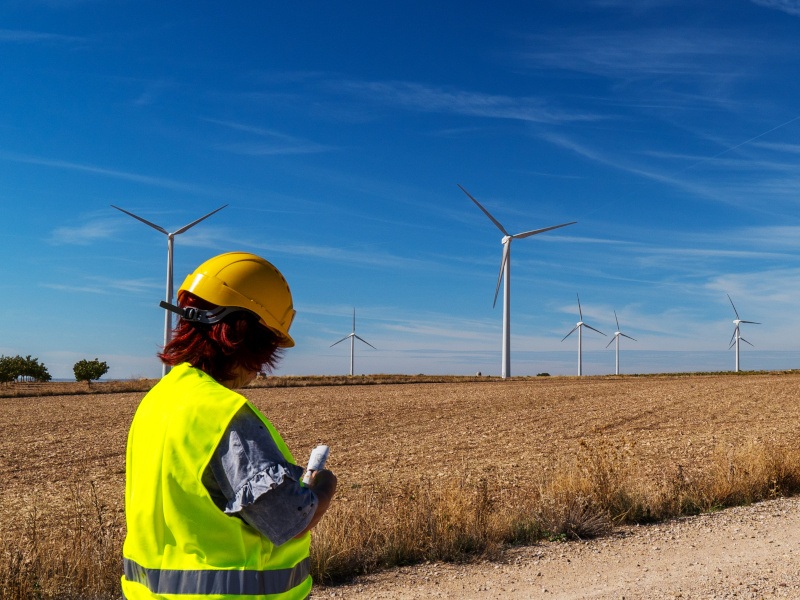 A woman dressed in a high-vis jacket and a hard hat stands in front of a field while turbines turn in the distance. It is a sunny day.