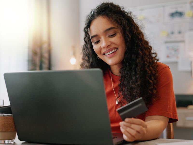 A young woman smiles as she looks at a credit card. She is sat at a desk and is on a laptop. The sun appears to be shining through a nearby window.