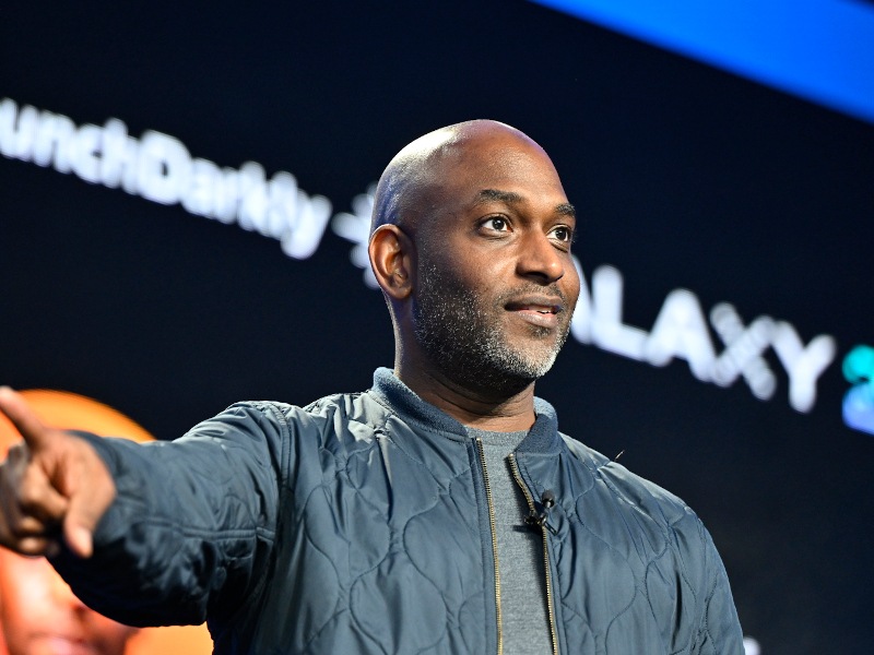 A man stands on stage pointing to the side with a large screen behind him.