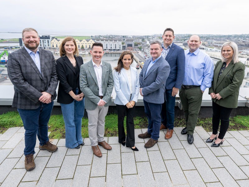 A group of eight people dressed in professional attire pose for a photograph on top of a building. A dockland can be seen in the background.